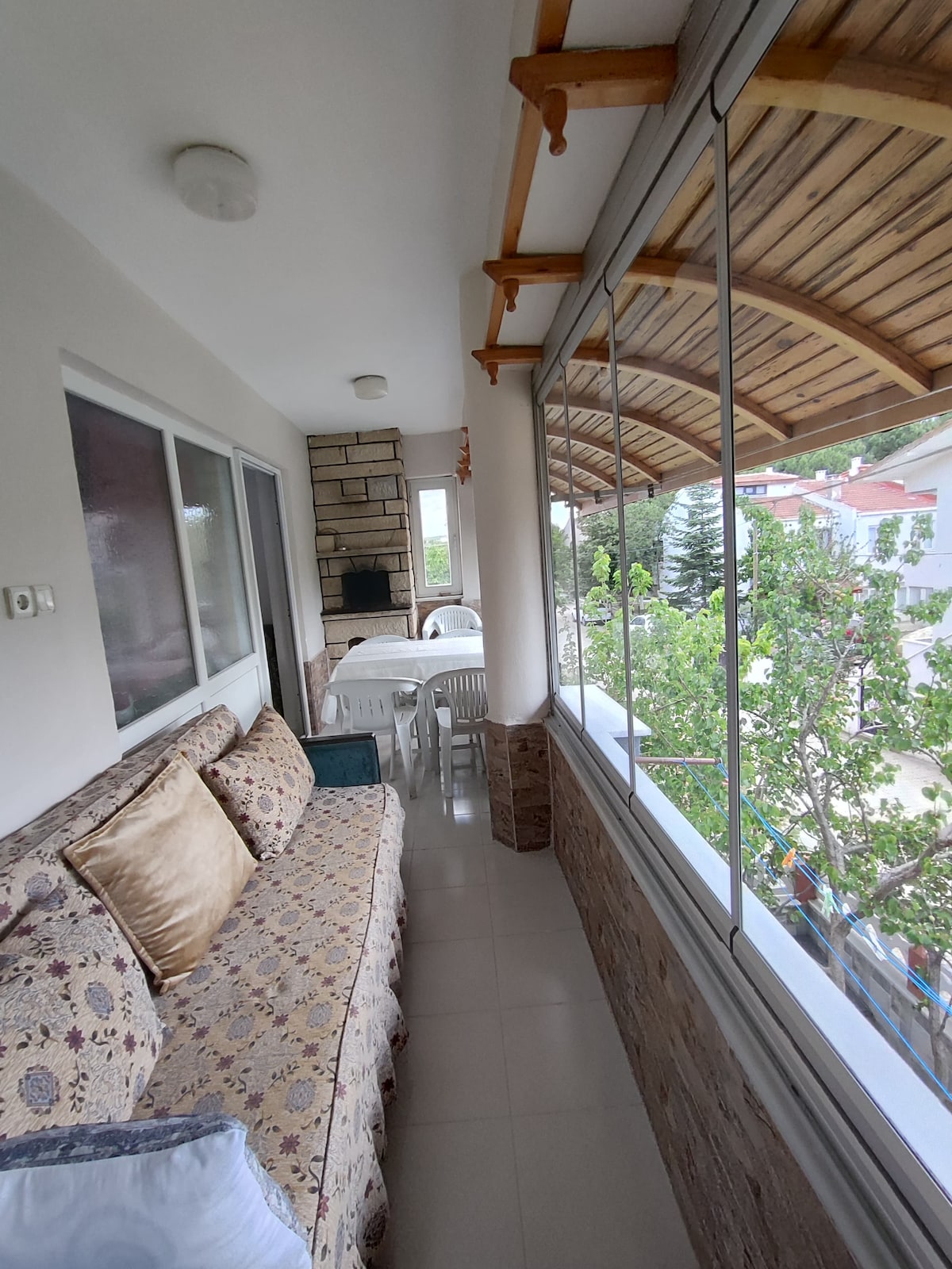 A sunlit balcony is furnished with a patterned sofa and a glass dining table. Light-colored walls are complemented by wooden beams overhead. A view of greenery is visible through large glass windows, creating a spacious and airy environment.