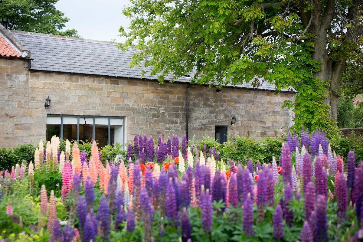Colorful flowers bloom in the foreground, featuring various shades of purple, pink, and orange. A stone building with several windows is visible in the background, partially shaded by lush greenery and trees.