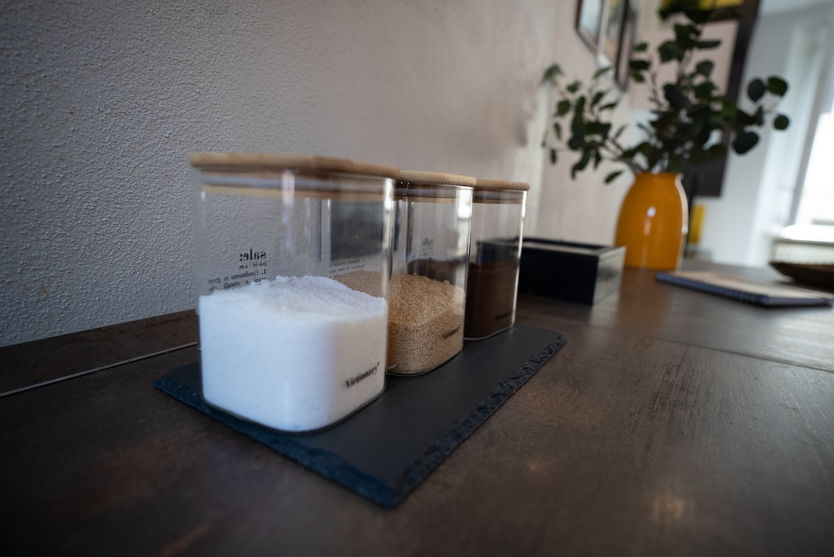 A clean countertop features three glass containers with wooden lids, filled with white sugar, brown sugar, and ground coffee. The containers are arranged on a dark slate tray, enhancing the modern aesthetic of the space. Natural light softly illuminates the area.