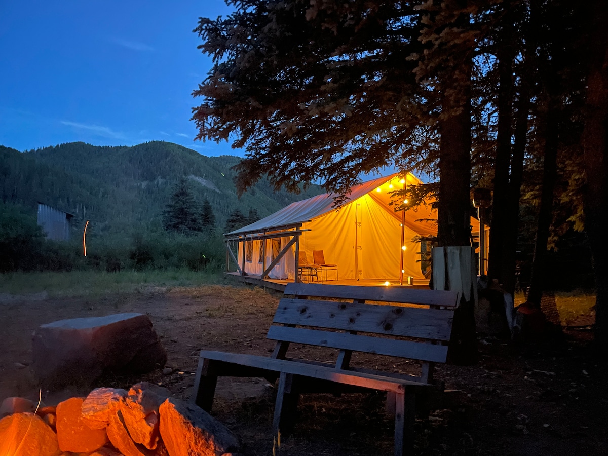 The tent is illuminated with warm lighting as dusk sets in, nestled among trees. A wooden bench is positioned in the foreground, facing a small fire pit with stones arranged around it. The surrounding mountains create a serene backdrop.