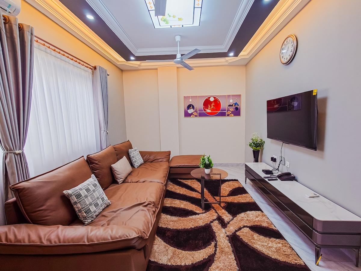 A spacious living area features a large, comfortable sofa adorned with decorative cushions. A sleek coffee table sits on a patterned area rug. Natural light filters through sheer curtains, while a wall-mounted flat-screen TV and a ceiling fan add modern convenience.