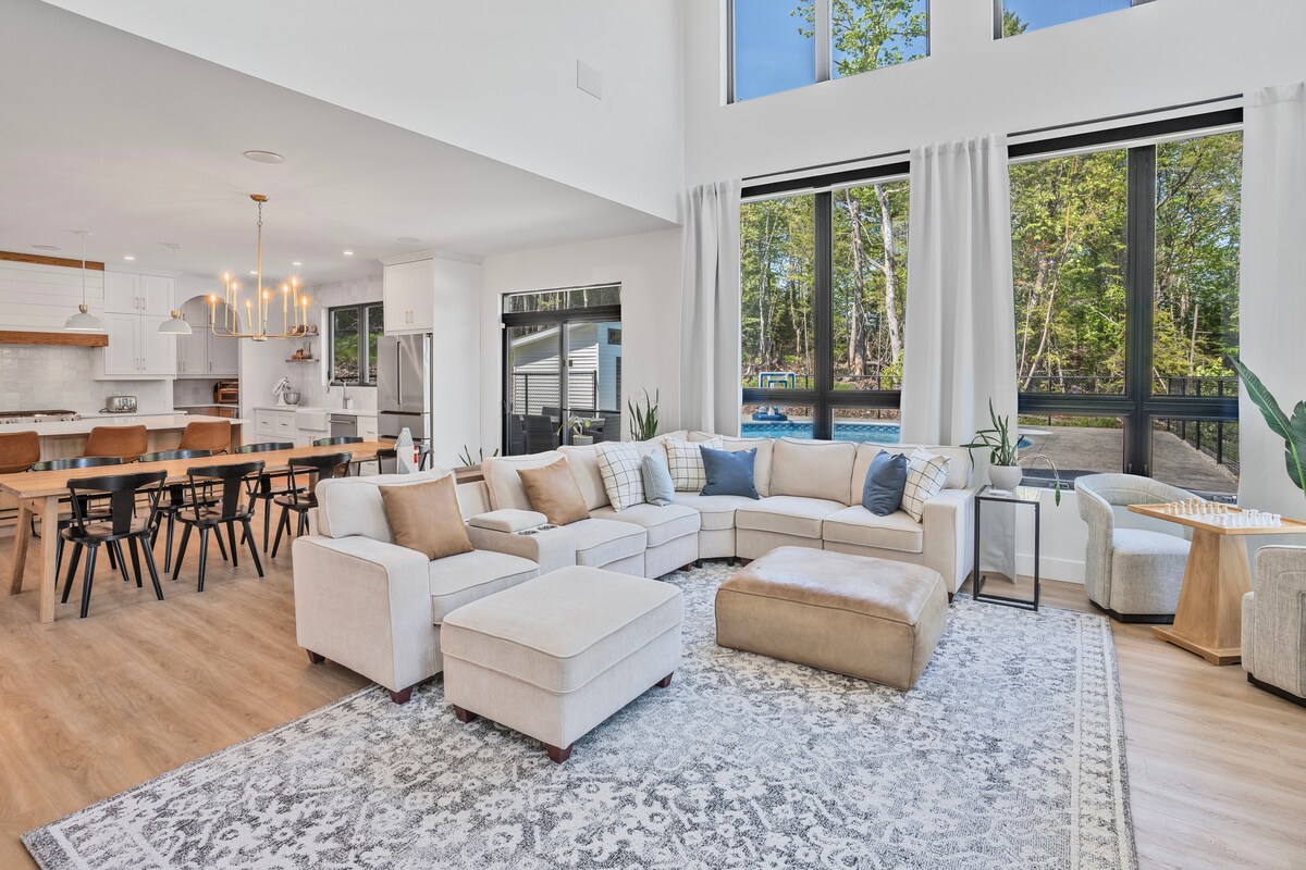 An open-concept living space features large windows that allow natural light to fill the room, providing views of the surrounding trees. The seating area is arranged around a light-toned area rug, with a spacious sectional sofa and a coffee table nearby. A dining table for multiple guests is visible in the background.