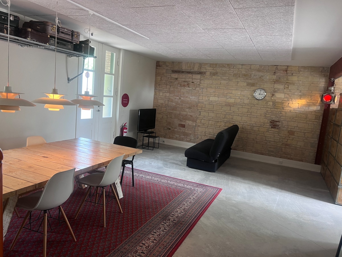 An open living space is presented with exposed brick walls and a large wooden dining table surrounded by light-colored chairs. A sleek black sofa is positioned nearby, with a television mounted on a wall. An antique clock hangs above an area rug that adds texture to the floor.
