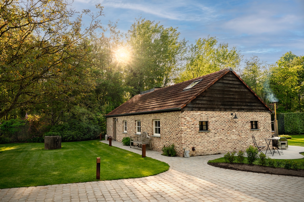 A charming single-story building is surrounded by greenery, featuring a natural stone façade and a sloped roof. Sunlight filters through the trees, casting a warm glow on the well-maintained lawn and the paving stones leading to an outdoor seating area.