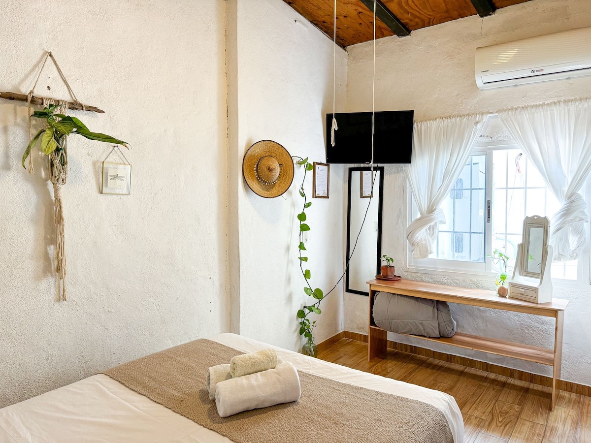 A simple bedroom setting is presented, featuring a spacious bed with neutral bedding and two rolled towels. A window with light, flowing curtains allows natural light in, while a wooden bench and hanging plant add a touch of greenery. A television is mounted on the wall.