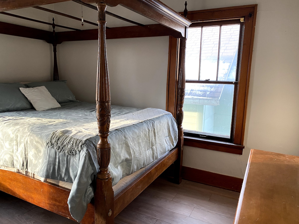 A spacious room features a four-poster bed with a light blue coverlet and decorative pillows. Natural light filters through a window with wooden trim, illuminating the room's warm wooden accents and flooring. A simple wooden dresser is positioned against the opposite wall.