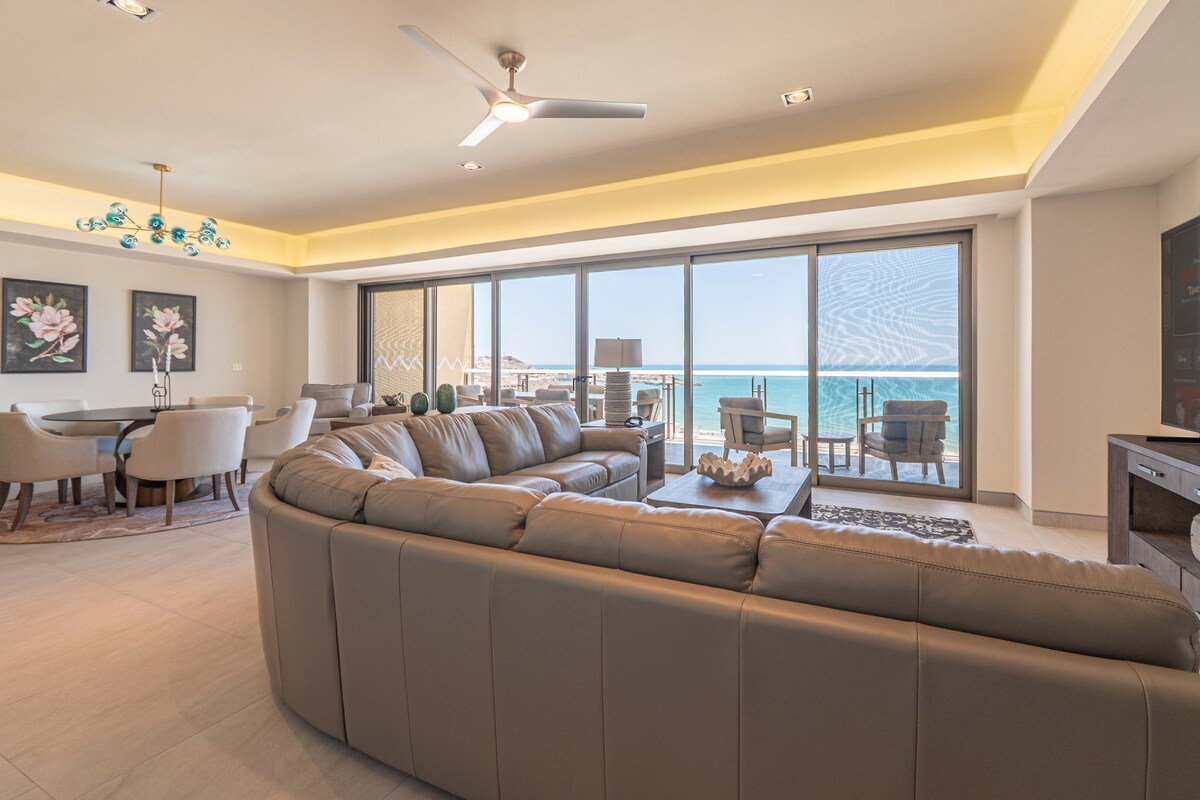 A modern living space features a spacious sectional sofa facing a large window with ocean views. Natural light enters the room, highlighting the stylish furnishings, including a wooden entertainment unit and a dining area with plush chairs. Elegant artwork adorns the walls.