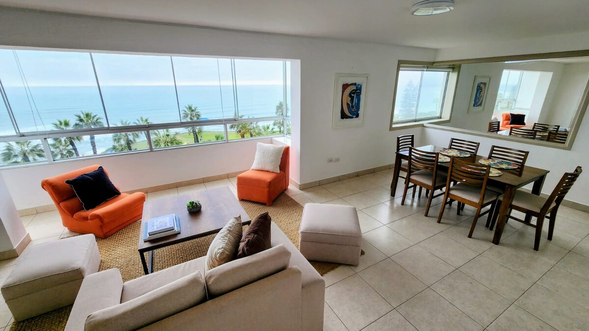 A spacious living and dining area features large windows offering a view of the ocean and palm trees. Two orange armchairs and a soft sofa create a comfortable seating arrangement. A wooden dining table is set for six, accompanied by neutral-toned ottomans on a light tiled floor.