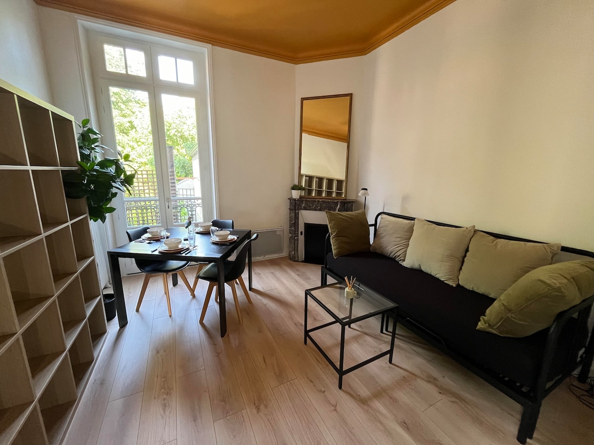 A living area is showcased, featuring a black sofa adorned with beige cushions. A dining table is set with dishes, surrounded by wooden chairs. Natural light streams through glass doors leading to an outdoor space, while a mirror reflects the warm tones of the ceiling.