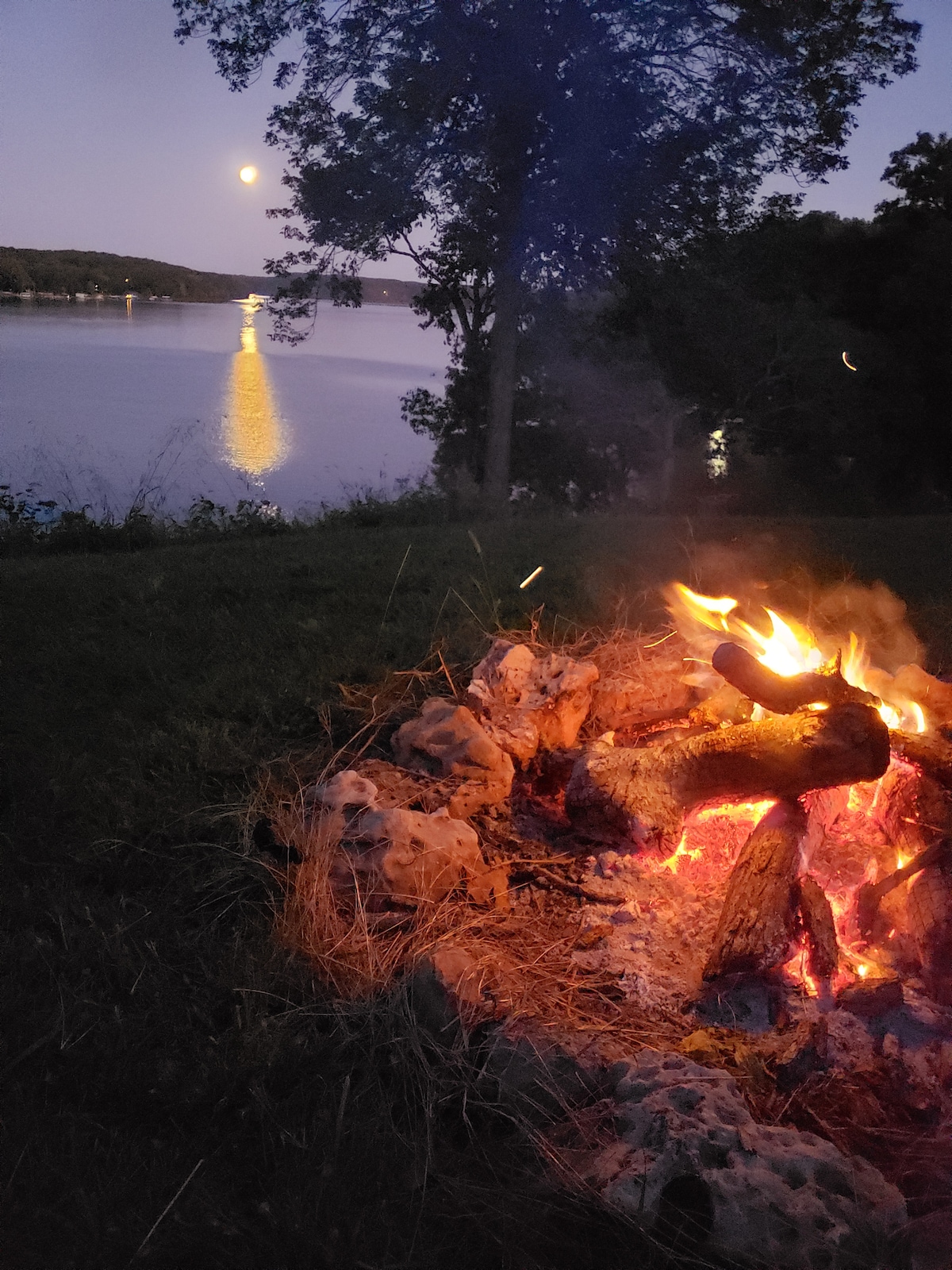 A crackling fire sits on a stone border, casting a warm glow against the darkening sky. The lake reflects the moonlight, creating a shimmering path on the water's surface. Trees frame the scene, enhancing the tranquil evening atmosphere.