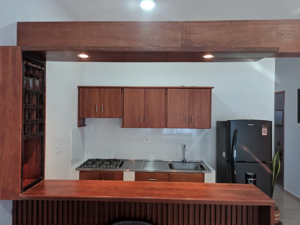 A modern kitchen area is displayed, featuring wooden cabinetry and a stainless steel sink. A gas stove sits adjacent to the counter, and a black refrigerator stands in the background. Soft ceiling lights illuminate the space, highlighting the warm wood tones.
