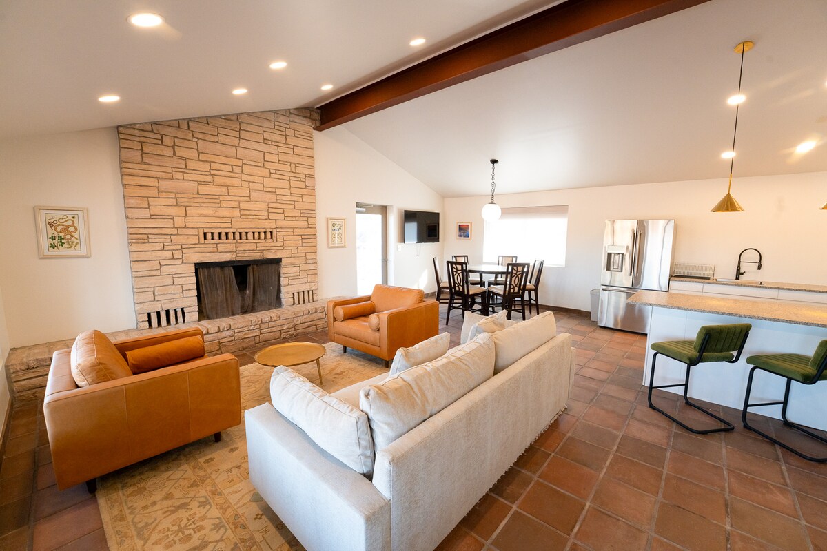 A spacious living area features a stone fireplace at one end, surrounded by two brown leather chairs and a cream-colored sofa. A dining table is set nearby, with a modern kitchen visible in the background, showcasing stainless steel appliances and an open layout.