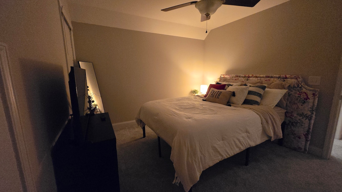 A cozy bedroom is seen, featuring a spacious bed with a floral headboard and decorative pillows. Soft lighting creates a calm ambiance, complemented by a ceiling fan. An entertainment unit is positioned against one wall, and a mirror stands nearby, reflecting the room's warmth.