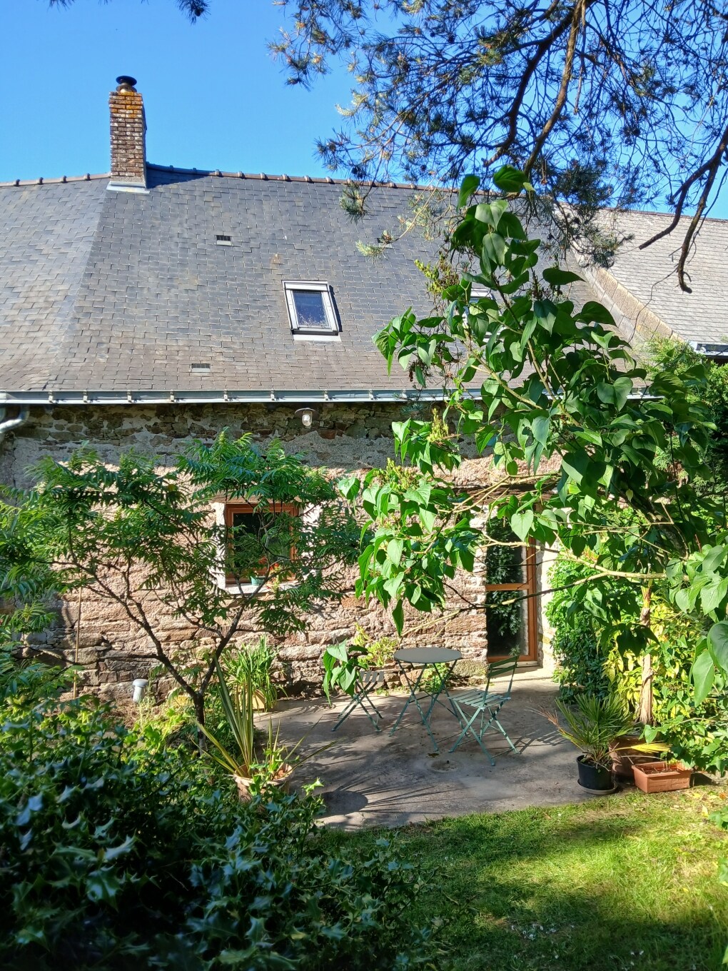 The exterior of a charming stone house is framed by greenery, showcasing a small private terrace with metal bistro-style chairs and a table. Sunlight filters through the trees, highlighting the rustic texture of the building and the inviting outdoor space.