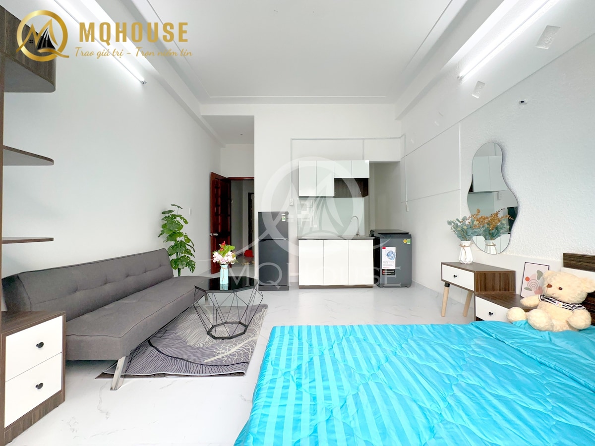 A modern living space features a gray sofa against a bright wall, accompanied by a minimalist coffee table. A blue bedspread covers the spacious bed, while a round mirror adds depth. A small kitchenette is visible in the background, contributing to the functional layout.