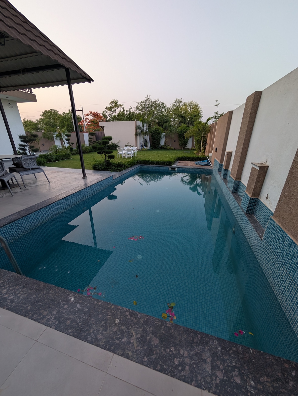 A private swimming pool features clear water adorned with floating flowers, surrounded by well-maintained greenery. The pool's tiled edges contrast with the smooth surface, and a spacious patio is visible beside it, complete with outdoor furniture for relaxation.