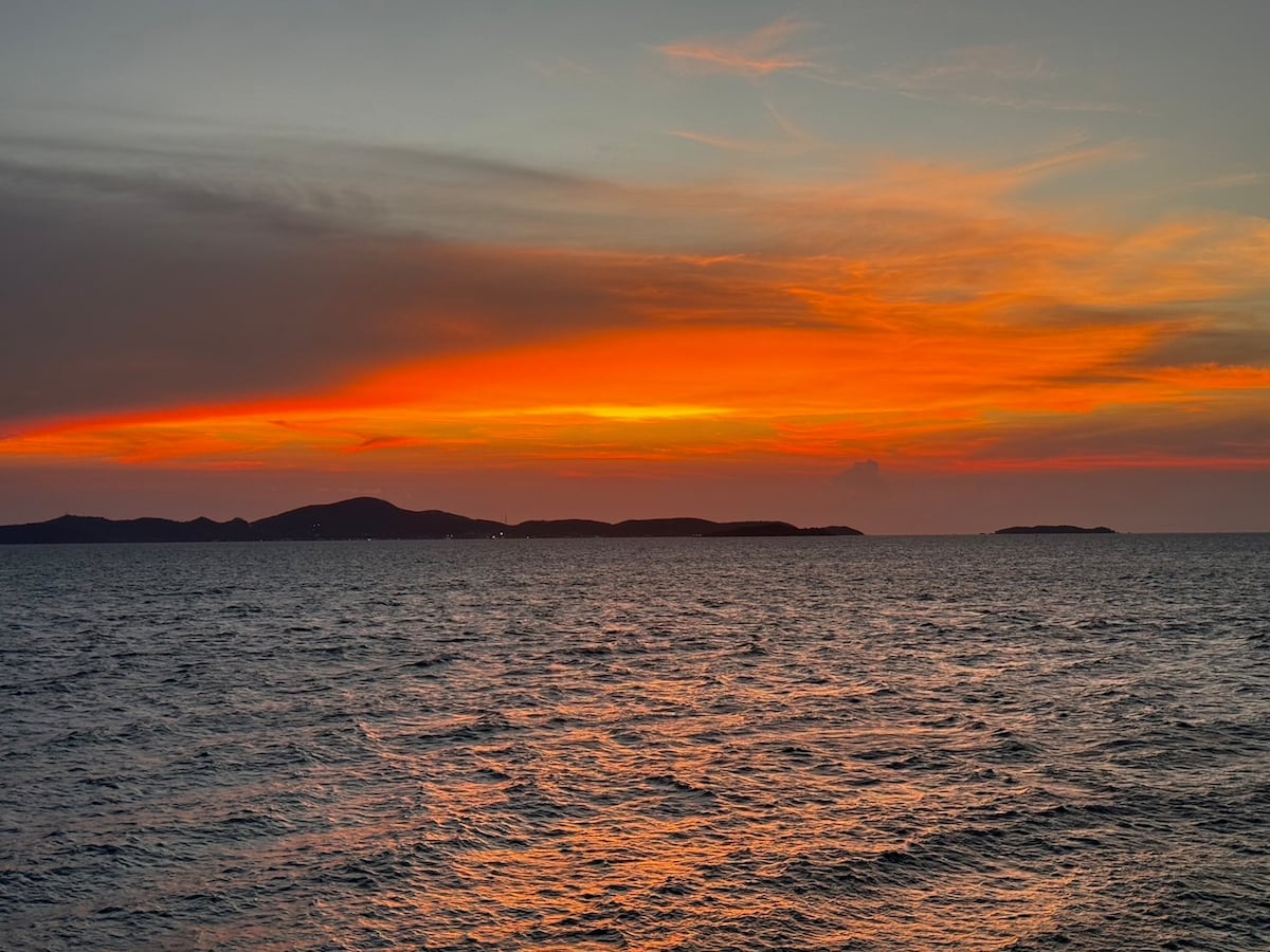 A serene seascape captures the expansive view of gentle waves reflecting the vibrant hues of a sunset. Layers of orange and pink paint the sky, blending into the distant silhouette of hills along the horizon, creating a calm and tranquil atmosphere.
