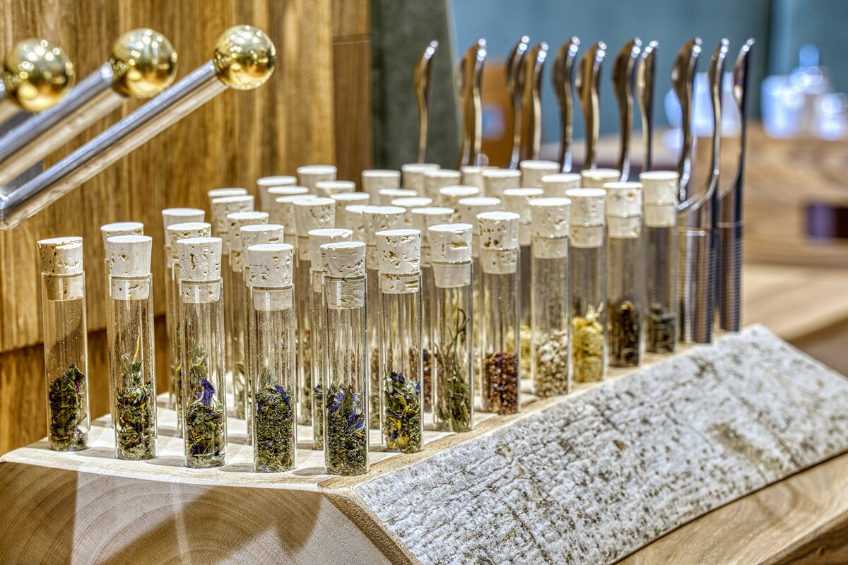 A wooden display holds a collection of glass vials containing various herbs and spices. Each vial features a cork stopper and is arranged neatly on a textured wooden base. The background is softly blurred, emphasizing the variety of natural ingredients.