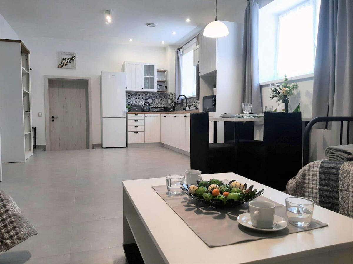 An open-concept living area is visible, featuring a dining table set with glassware and decorative items. A modern kitchen equipped with appliances is seen in the background. Natural light enters through a window, illuminating the neutral-toned decor and sleek cabinetry.