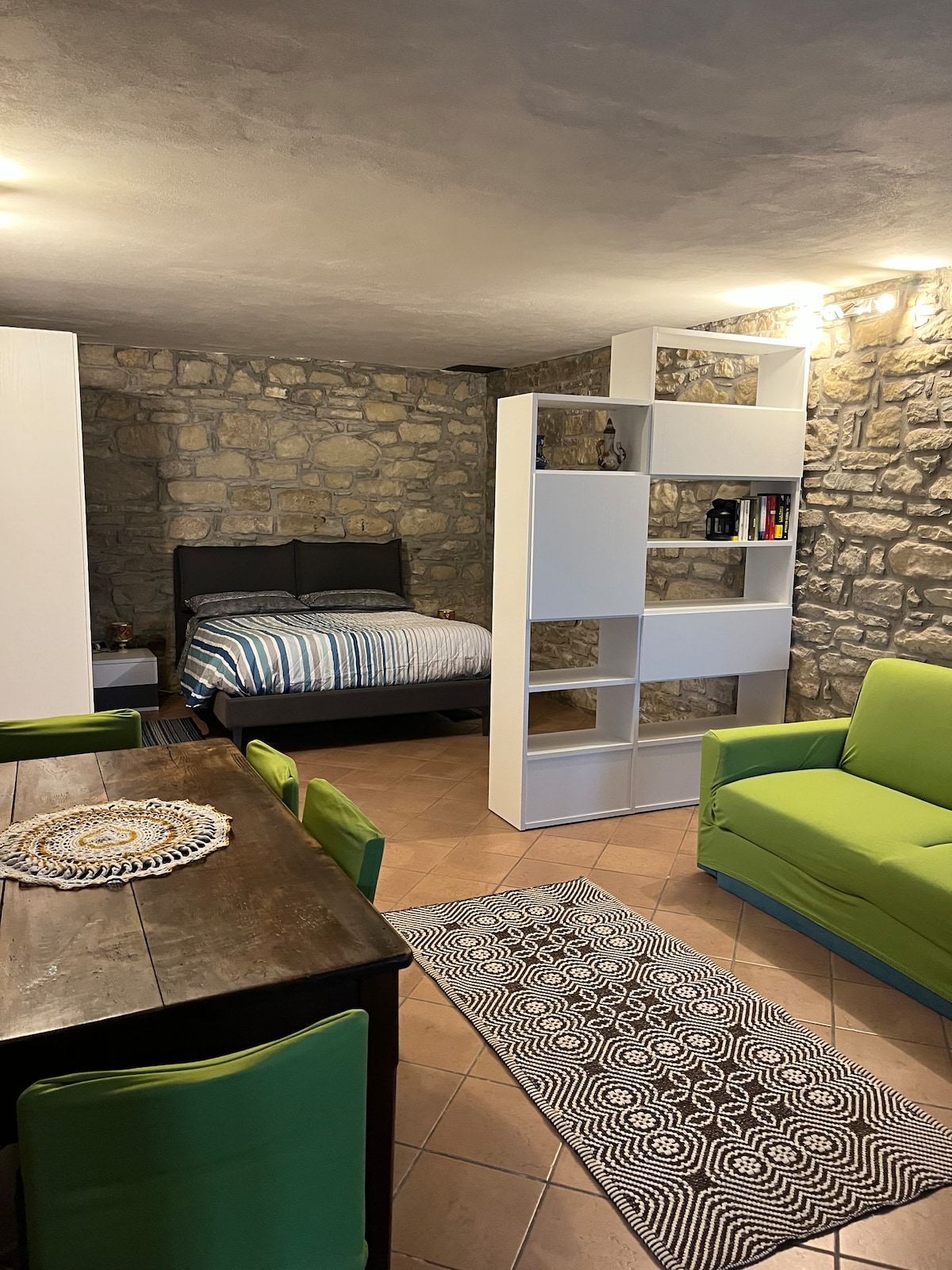 A cozy living space features a spacious bed with striped bedding, a white bookshelf displaying various items, and a dining table for four. A green sofa adds a pop of color, while the stone wall provides a rustic backdrop. Natural light is provided by overhead fixtures.