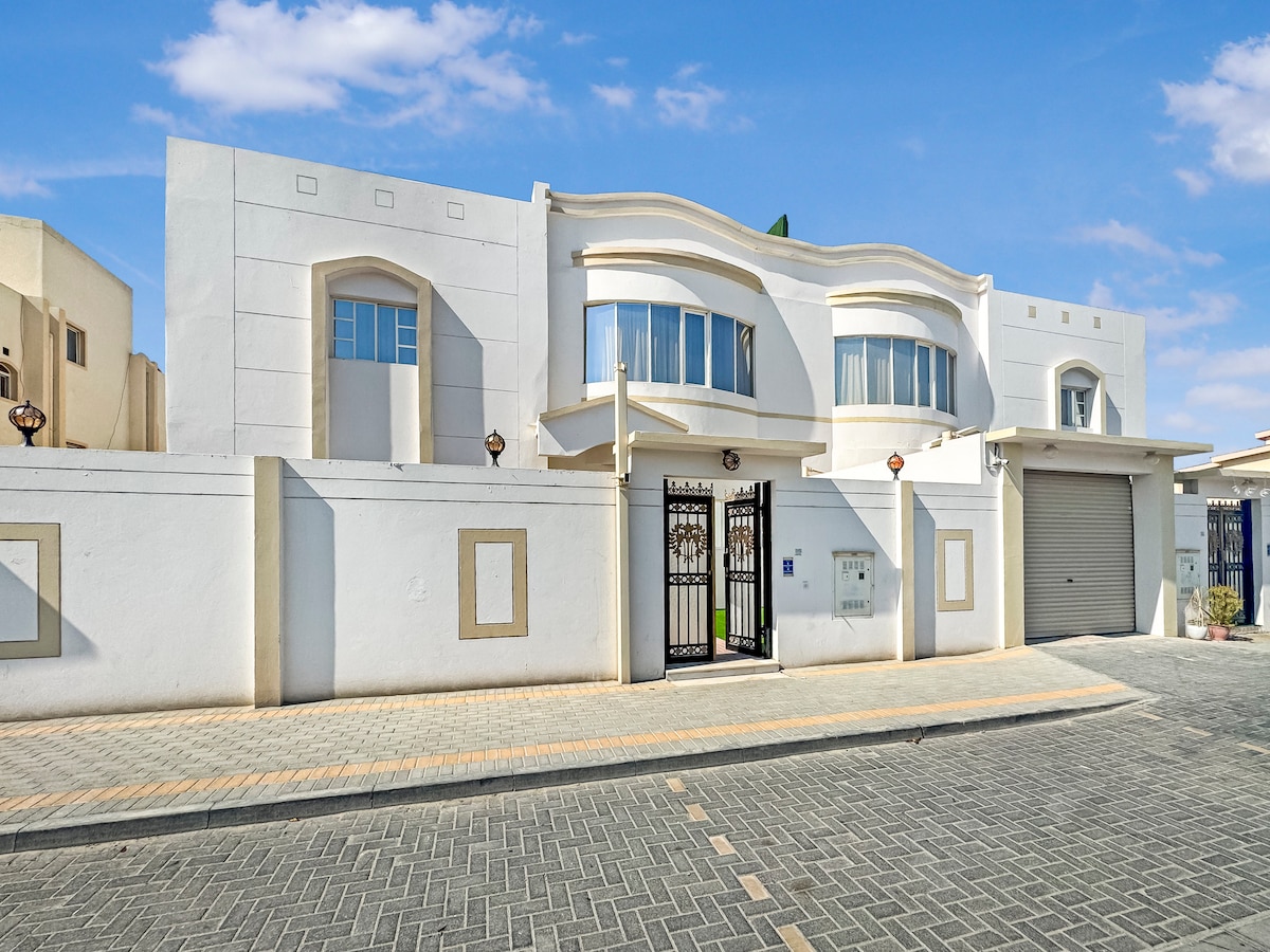 The exterior of a spacious villa is presented, featuring a modern white facade with arched windows and a decorative entrance gate. A well-maintained garden area is visible in front of the property, while a paved path leads to the entrance along the surrounding block wall.