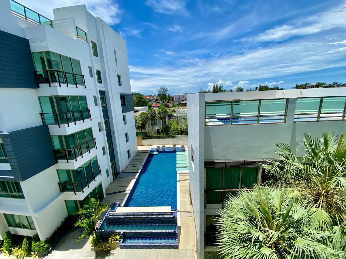 A spacious outdoor pool is framed by palm trees and modern buildings, providing a serene view. The pool area features sun loungers and a shallow edge, while the surrounding architecture showcases clean lines and large windows that allow for abundant natural light.
