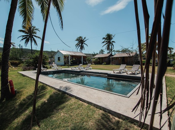 A serene outdoor area features a rectangular swimming pool surrounded by a sun deck equipped with lounge chairs. Lush greenery and palm trees provide a tropical setting, while a charming white building and wooden structures are visible in the background.