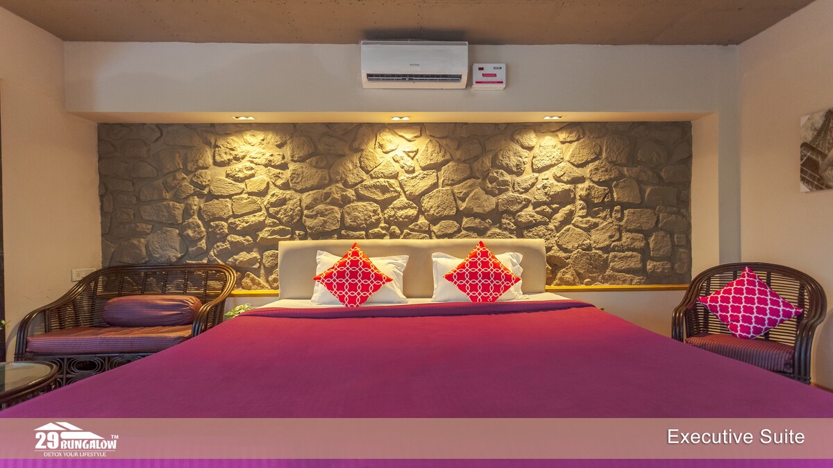 A spacious suite features a large bed with decorative pink cushions against a textured stone wall. The room is illuminated by soft lighting, with air conditioning visible above. Two wicker chairs with patterned cushions complement the inviting decor.