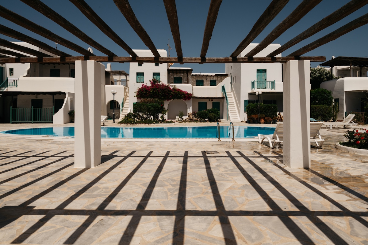 The outdoor area features a shared swimming pool surrounded by sun loungers. Wooden beams create a structured pattern overhead, casting shadows on the stone patio. Lush greenery and colorful flowering plants can be seen in the background, adding to the inviting ambiance of the space.