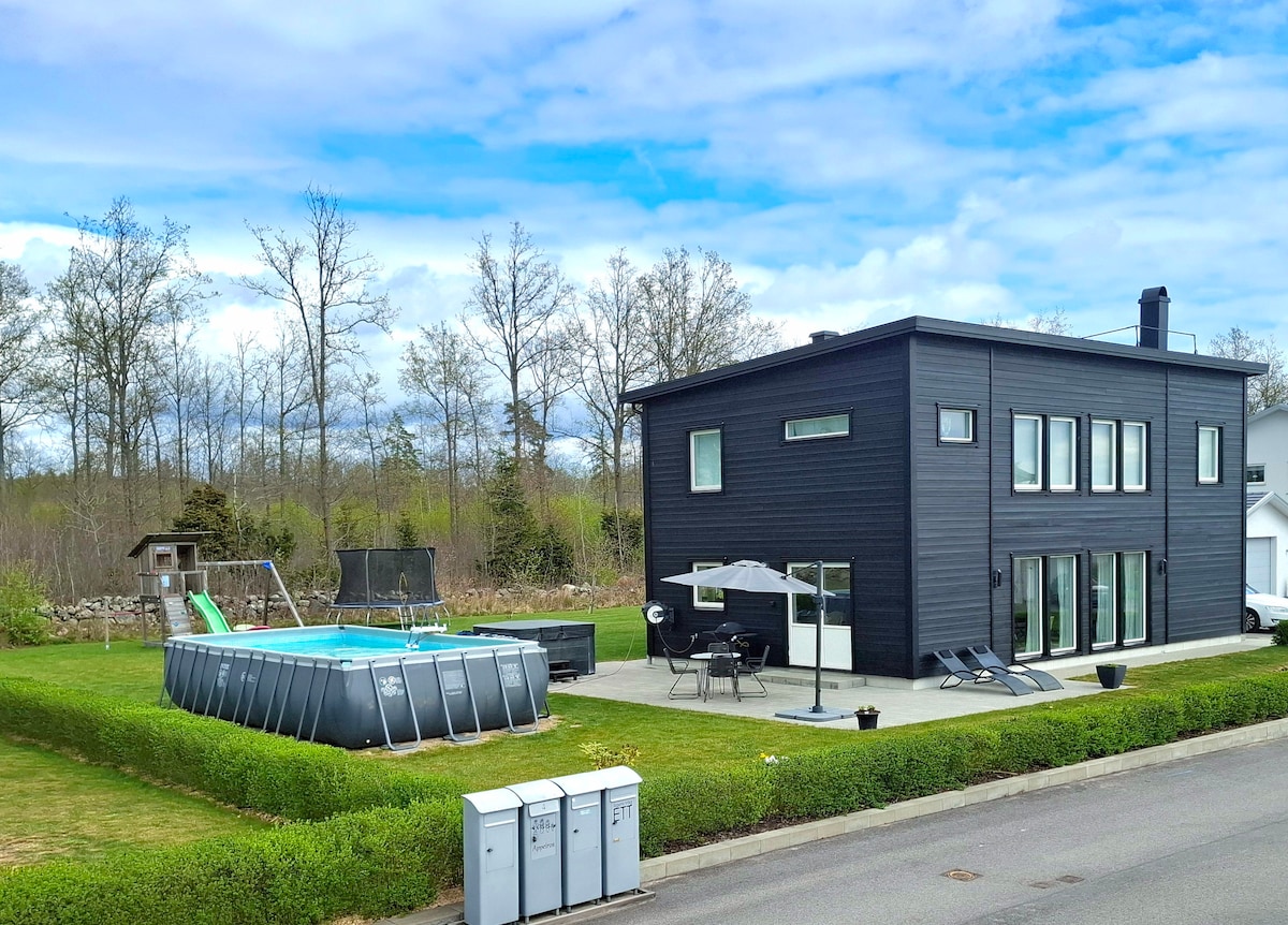 A modern, two-story villa is showcased, featuring a spacious outdoor area with a pool, surrounded by greenery. The exterior includes large windows and a sleek black facade. Lounge chairs and a dining set are arranged on the patio, offering an inviting space for relaxation.