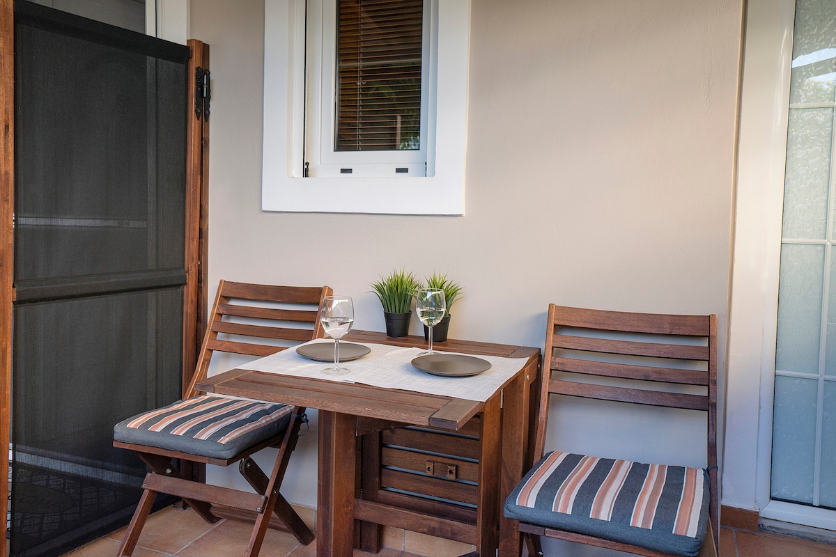 A small outdoor dining area features a wooden table set for two, with two folding chairs adorned with striped cushions. A potted plant adds a touch of greenery to the table, enhancing the inviting atmosphere of the space.