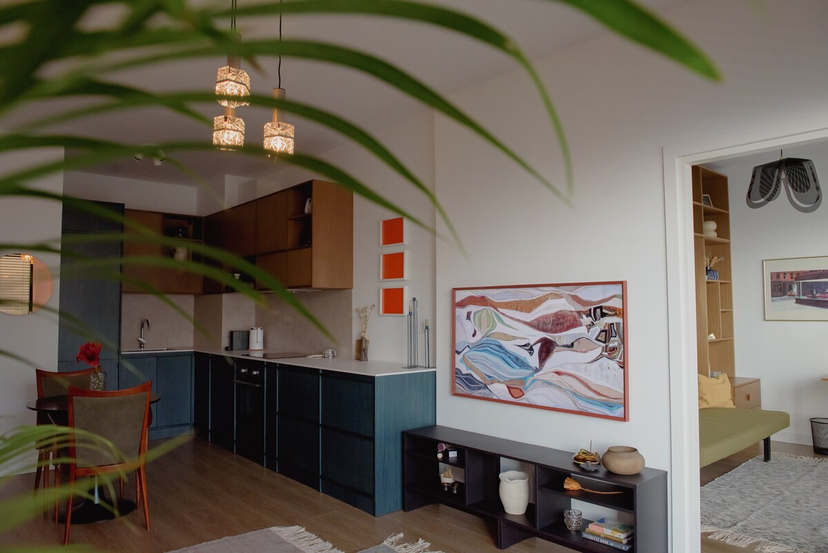 The image captures a contemporary living space characterized by clean lines and handpicked furnishings. A modern kitchen is visible, featuring wood cabinetry and subtle lighting. An artistic wall piece adds interest, while greenery in the foreground enhances the inviting ambiance of the area.