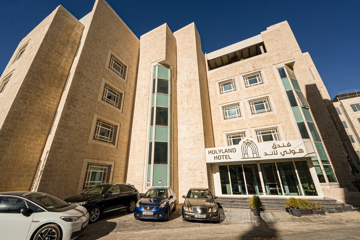 The hotel exterior is characterized by a modern design with large glass windows and a neutral stone facade. Several vehicles are parked in front of the building, and the hotel name is prominently displayed on a sign.