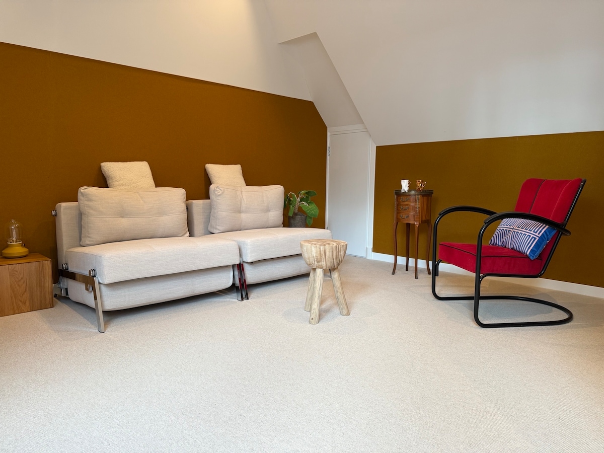 A cozy living space is furnished with a light-colored sofa and an armchair in bold red. A small, natural wood stool and a decorative side table enhance the setting, while a potted plant adds a touch of greenery. The walls are painted in a warm brown tone.