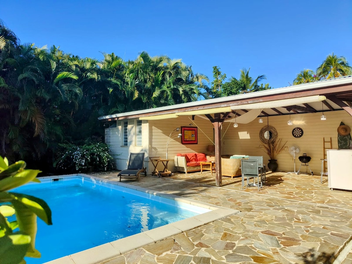 A tranquil outdoor area features a rectangular pool surrounded by a stone patio. A covered seating area with comfortable seating is visible, along with a dining space. Lush greenery and tropical plants provide privacy and enhance the serene setting.