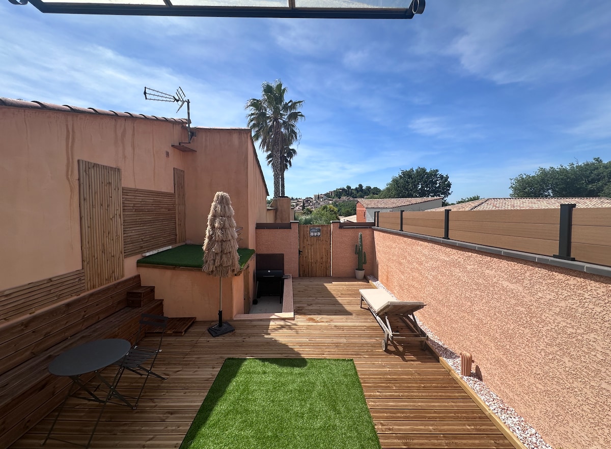 An outdoor terrace features wooden decking with a sunbed positioned alongside a patch of green grass. A small black table and chairs provide a seating area, while a large umbrella offers shade. A tall palm tree and a structured fence complete the serene setting.