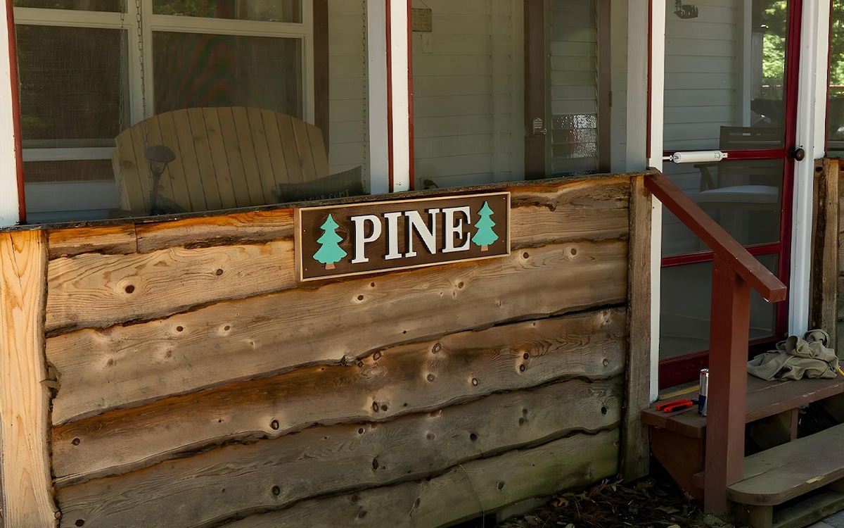 The exterior of the cabin features a wooden wall with a rustic sign displaying the word 'PINE,' accompanied by illustrations of pine trees. A screened-in porch area is partially visible, with a comfortable chair positioned nearby.