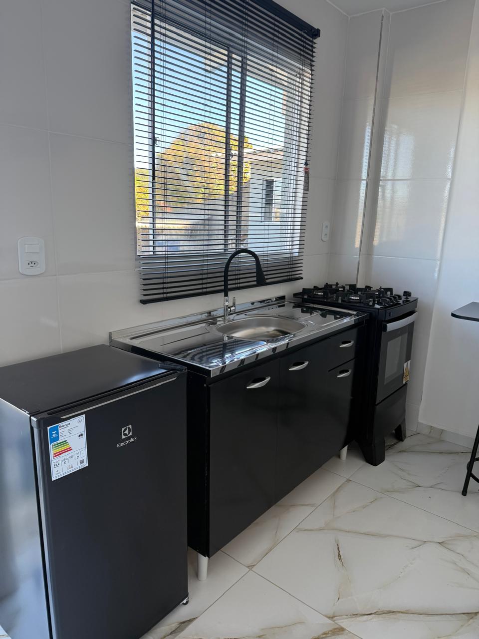 The kitchen features a compact layout equipped with a black refrigerator, a stainless steel sink, and a gas stove. Natural light enters through a large window adorned with horizontal blinds, providing a bright atmosphere. Sleek cabinetry complements the modern design.
