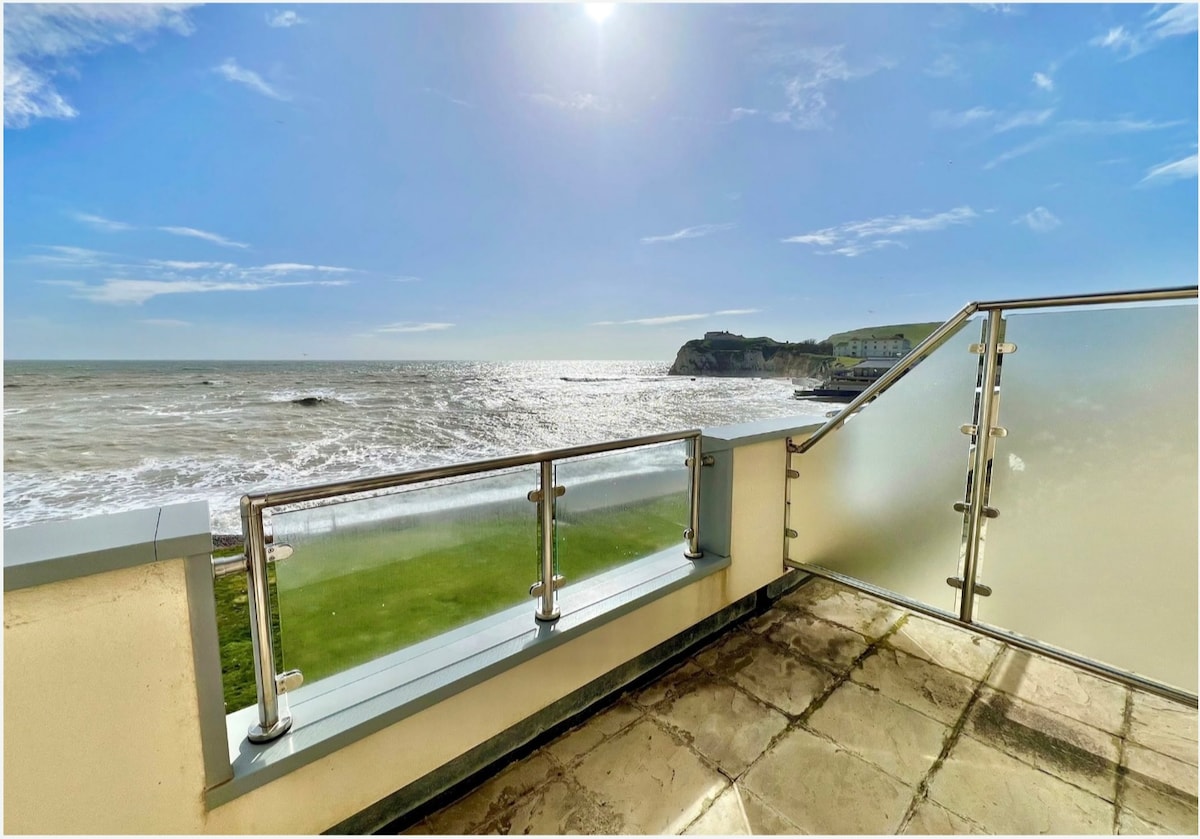 A private balcony offers unobstructed views of the sea and coastline. The glass railing enhances the panoramic perspective of the waves and sky, with sunlight reflecting off the water. The outdoor flooring consists of large stone tiles, providing a smooth surface.