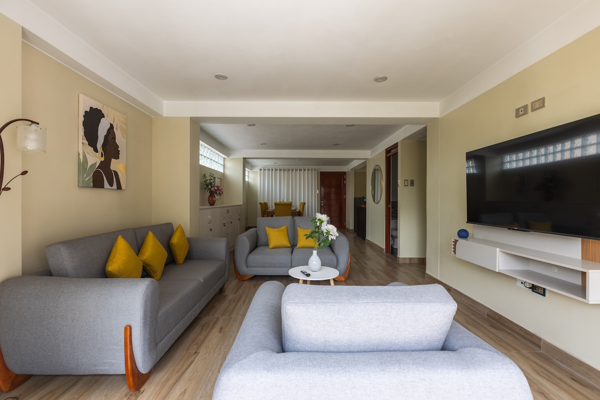 The modern living area features a spacious layout with two gray sofas accented by yellow cushions. A low coffee table is positioned in front of the seating. A large flat-screen TV is mounted on the wall, while natural light enters through the windows, creating a welcoming environment.
