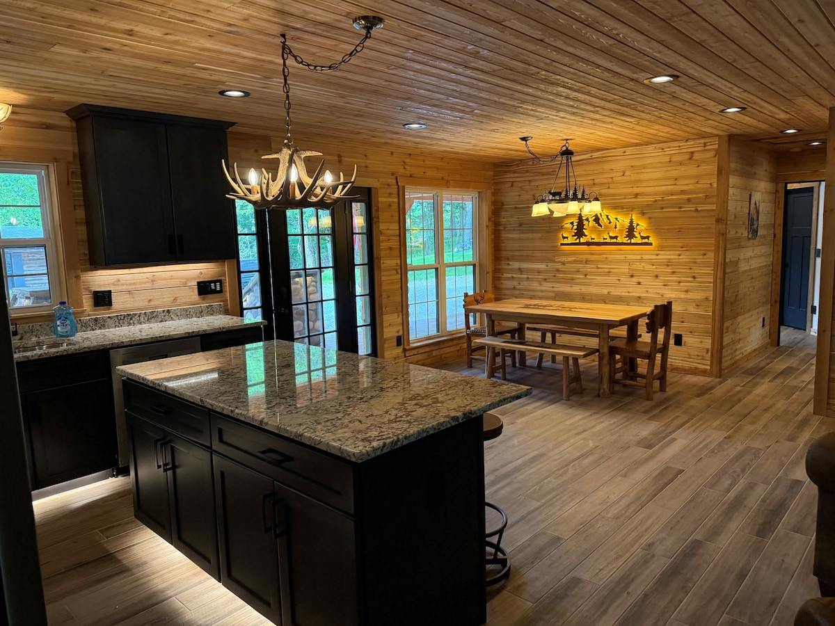 An open-concept living area is presented, featuring a kitchen with a dark cabinetry and a granite island, illuminated by a rustic chandelier. A wooden dining table is positioned against a wall of windows, while wooden paneling covers the walls, adding warmth to the space.