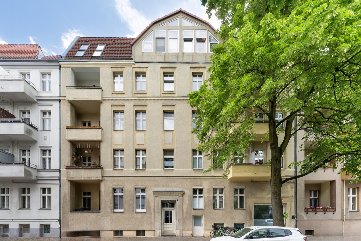 An exterior view of a spacious building featuring a symmetrical design with multiple windows. The façade is complemented by balconies and large trees, providing shade and greenery. The entryway is marked by a traditional door, with a well-maintained street visible in front.