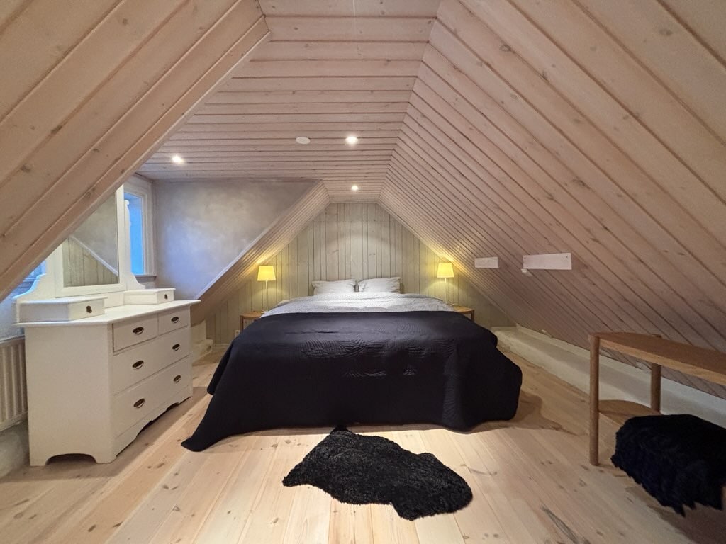 The lofted sleeping area features sloped wooden ceilings adorned with light wood paneling. A spacious bed is centered beneath a window, flanked by two lamps on nightstands. A soft black rug lies on the light wooden floor, complementing a white dresser and a simple wooden bench.