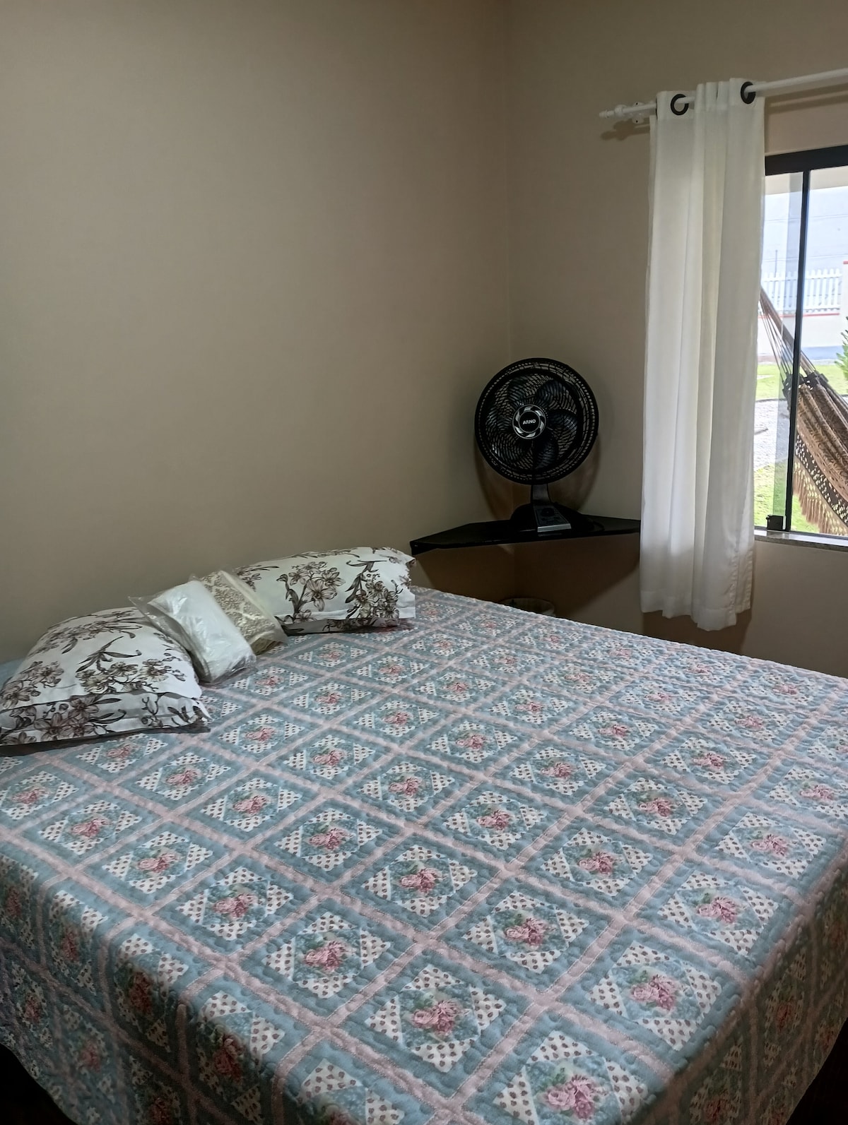 A cozy sleeping area features a spacious bed dressed in a patterned quilt. Decorative pillows are arranged at the head of the bed. A small fan is positioned on a side table, and natural light enters through a window adorned with light curtains.