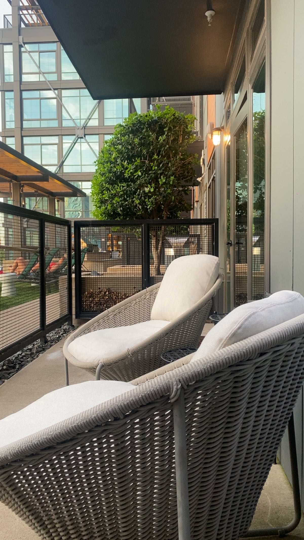 A small patio area is visible, featuring two woven chairs with plush white cushions. A lush green plant is situated nearby, and a clear view of the surrounding architecture is provided through glass panels. Natural light enhances the inviting outdoor space.