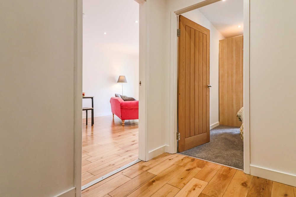 A view down a hallway showcases a blend of warm wooden flooring and soft carpeting. Light from recessed ceiling fixtures illuminates the inviting entrance area that leads to a living room with a red sofa and a bedside area with a partially visible door.