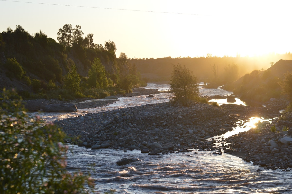 A serene river meanders through a landscape dotted with lush greenery. The sun casts a warm glow over the water, creating shimmering reflections. Smooth pebbles line the riverbank, and distant trees frame the scene, enhancing the sense of tranquility.
