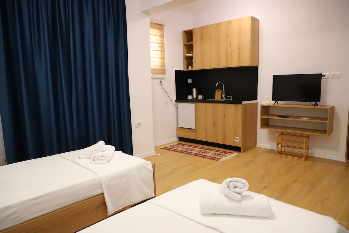 The image showcases a bright room featuring two single beds with white linens and neatly rolled towels. A small kitchenette with wooden cabinetry and a TV is visible in the background, complimented by a window covered with light-filtering blinds and deep blue curtains.