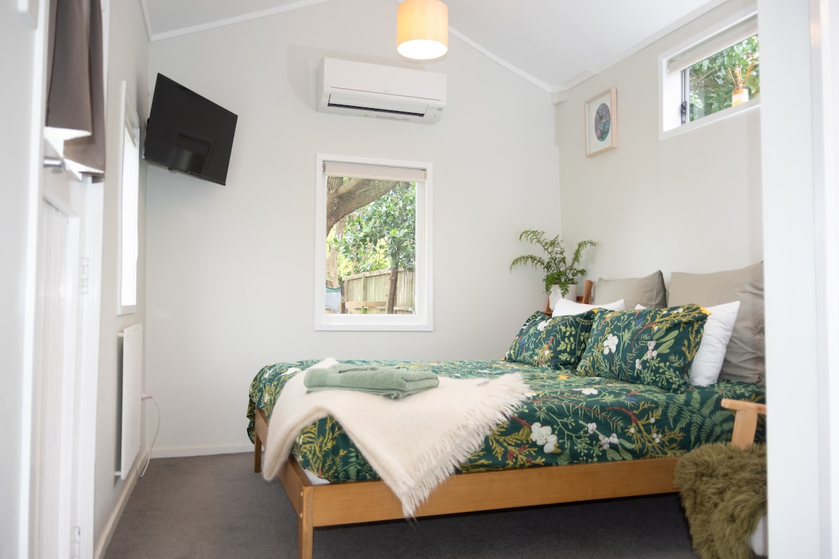 A cozy sleeping area features a wooden bed with a floral-patterned duvet and warm throws. Two windows provide natural light, framed by light curtains, and a small television is mounted on the wall. A small plant adds a touch of greenery to the space.