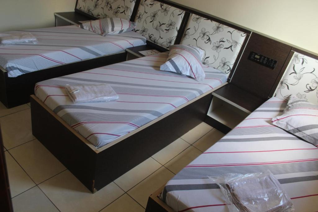 A room setup featuring four single beds arranged neatly within a spacious area. Each bed has a striped mattress with decorative headboards. White linens and towels are placed on some beds, and the flooring consists of light-colored tiles, creating a simple and functional space.